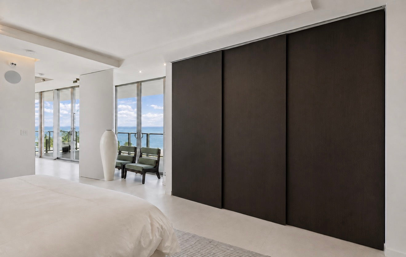 Three Espresso-stained maple Minimalist Slab doors on recessed ceiling tracks separating an ocean-view bedroom and bathroom.